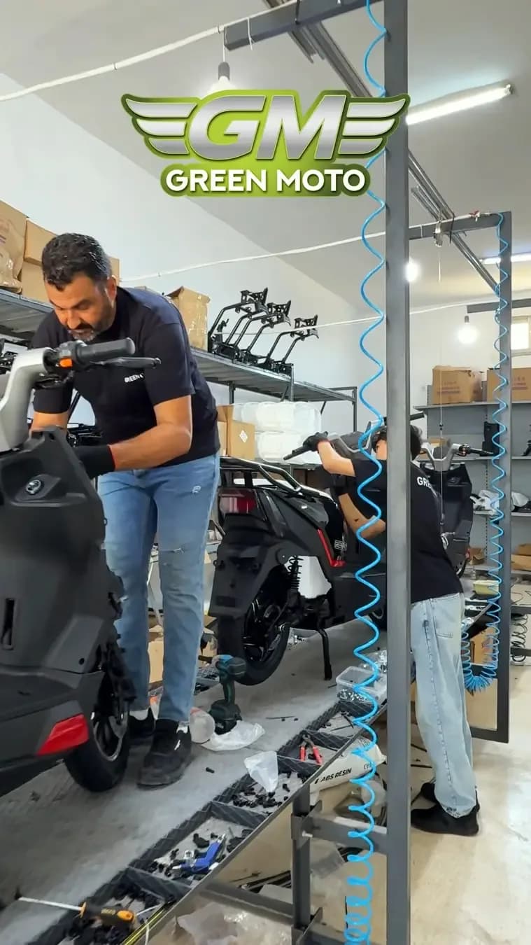 لقطة من فيديو عمودي: فنيّان بقميصين أسودين يجمّعان دراجات سكوتر في الورشة، تحت شعار Green Moto