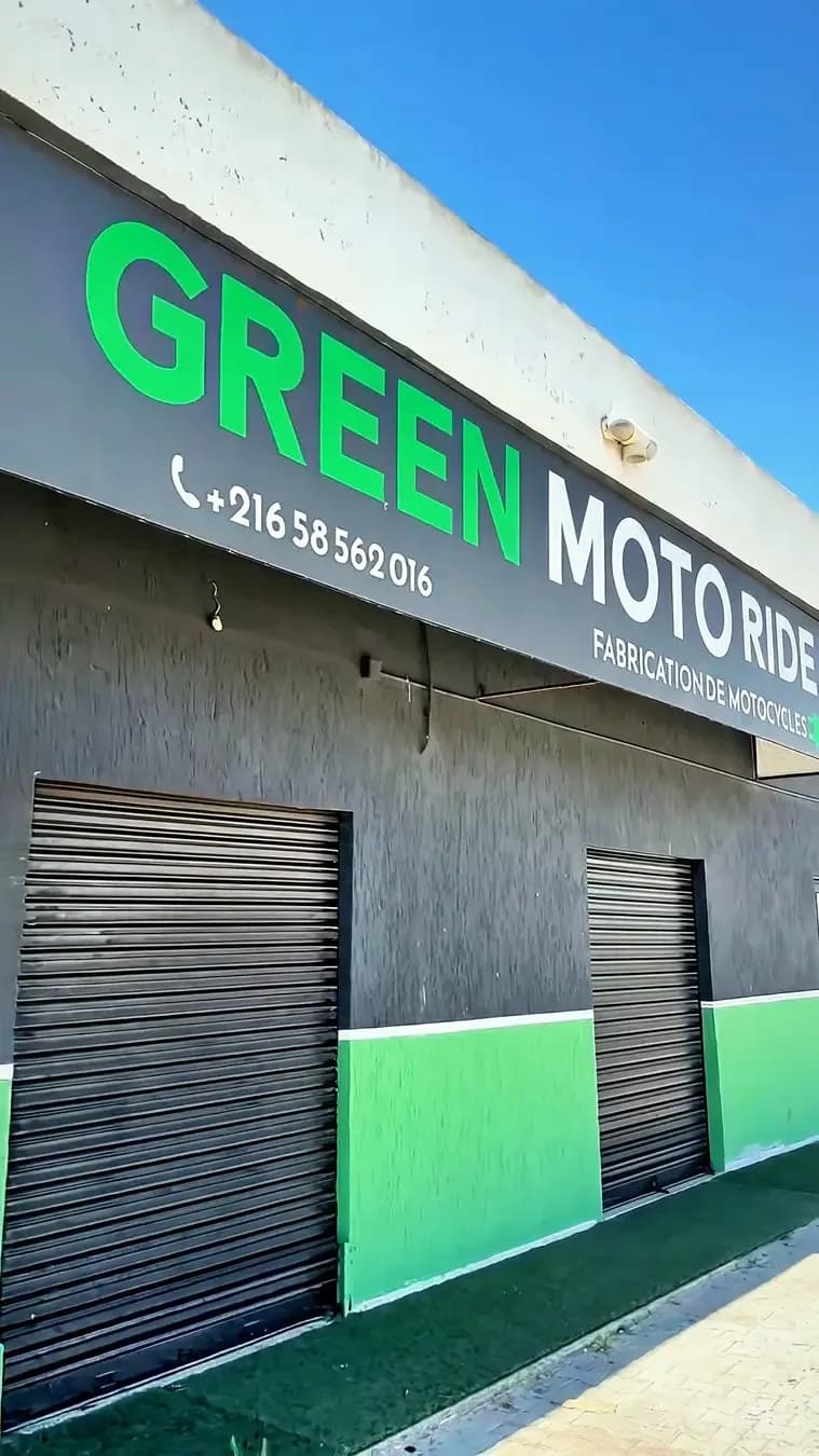 لقطة من فيديو عمودي: واجهة مطلية بالأخضر والرمادي الداكن، عليها لافتة «Green Moto Ride – Fabrication de motocycles» وستارتان معدنيتان
