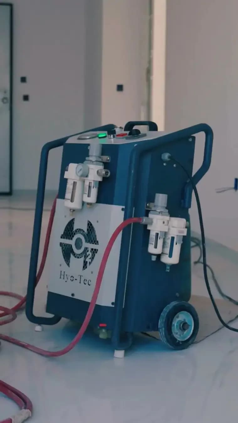 Still from a vertical video on the page: a navy-blue HyO-Tec machine on wheels, with a white panel bearing the brand’s logo, filter-regulators on its front and side, and a red hose, in a bright room